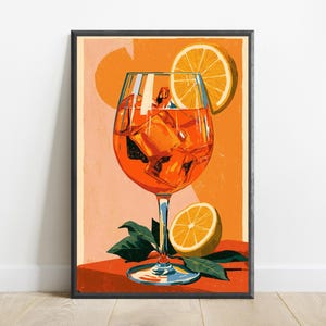 May include: A framed art print featuring a vibrant orange cocktail in a stemmed glass filled with ice cubes. An orange slice garnish rests on the rim, while another half-orange and green leaves sit at the base against a warm orange background.