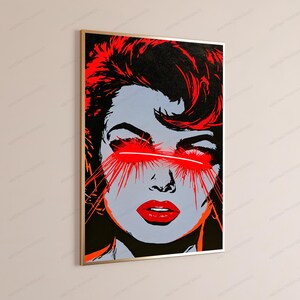 May include: A rectangular wall art print in a gold frame featuring a stylized portrait of a person with light gray skin, black hair, and bright red lips. A horizontal red beam of light crosses the eyes with radiating red lines.