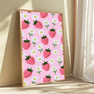 May include: A rectangular framed wall art piece featuring a repeating pattern of red strawberries with green stems and white daisies with yellow centers on a light pink background. The frame is made of light-colored wood with a thin profile.