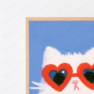 May include: A framed wall art print featuring a white cat wearing bright red heart-shaped sunglasses against a solid light blue background. The illustration has a textured, chalk-like appearance with a simple light wood frame border.