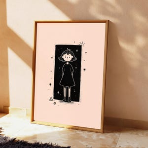 May include: A framed wall art print featuring a black and white line drawing of a person with short hair wearing a dress, centred within a black rectangular background on a light peach field, surrounded by small star and circular graphic accents.