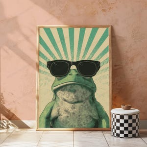 May include: A rectangular wall art print featuring a green frog wearing black sunglasses. The background consists of a light yellow and green sunburst pattern with a dotted texture. The frog is centered, facing forward, with a textured skin appearance.