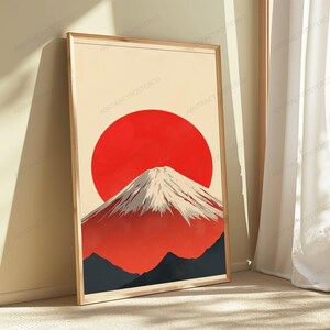 May include: A rectangular wall art print in a light wood frame featuring a graphic illustration of a snow-capped mountain peak set against a large, solid red circular sun on a cream-colored background, with dark gray foothills at the base.