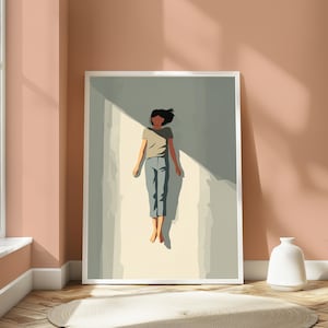 May include: A framed wall art print featuring a stylized illustration of a figure with dark hair wearing a beige t-shirt and blue pants, floating in a vertical shaft of light against a muted sage green background with sharp shadows.