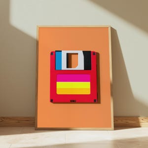 May include: A framed wall art print featuring a stylised floppy disk graphic centred on an orange background. The floppy disk is red with a blue and black top section, a white centre cutout, and a horizontal label with pink, yellow, and orange stripes.