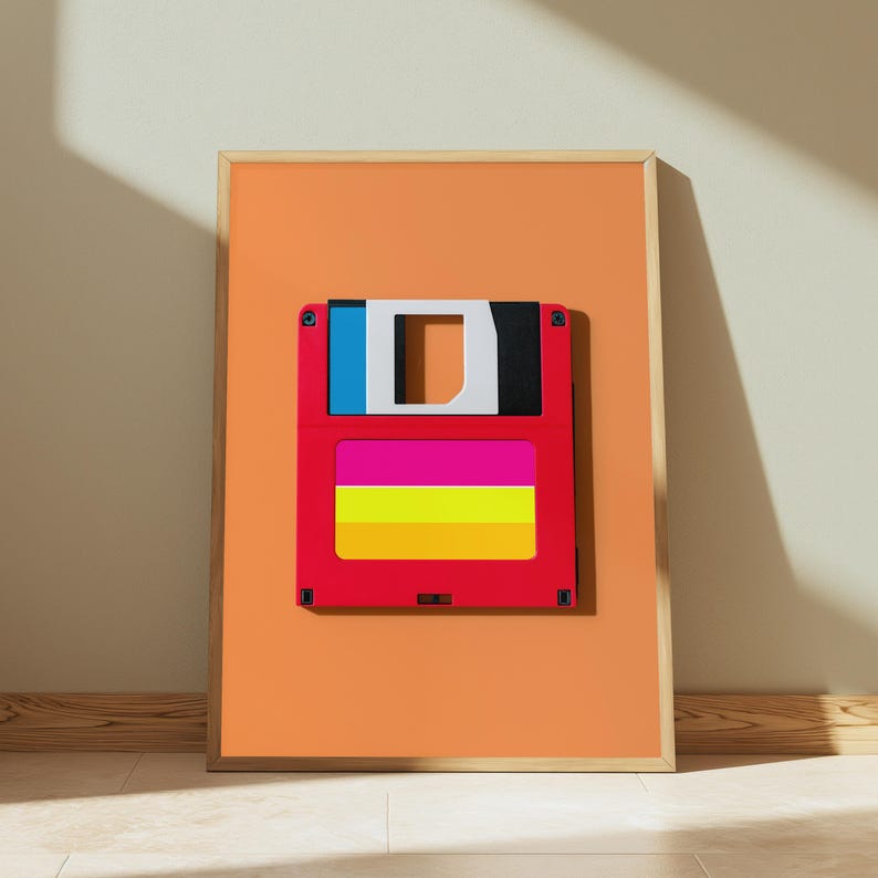 May include: A framed wall art print featuring a stylised floppy disk graphic centred on an orange background. The floppy disk is red with a blue and black top section, a white centre cutout, and a horizontal label with pink, yellow, and orange stripes.