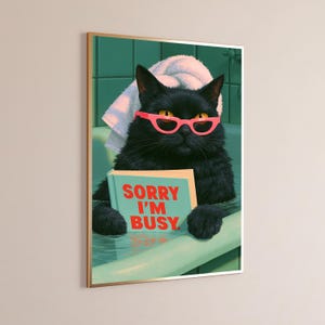May include: A framed wall art print featuring a black cat in a bathtub with a white towel on its head. The cat wears bright pink cat-eye glasses and holds a light blue book with the red text SORRY I'M BUSY printed on the front cover.