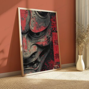 K&ouml;nnte beinhalten: Ein gerahmter vertikaler Wandkunstdruck mit einer detaillierten Graustufenillustration einer Hannya-Maske mit markanten H&ouml;rnern und scharfen Z&auml;hnen. Das Kunstwerk ist mit roten Farbspritzern akzentuiert und enth&auml;lt ein vertikales rotes rechteckiges Feld mit schwarzen japanischen Kanji-Schriftzeichen.