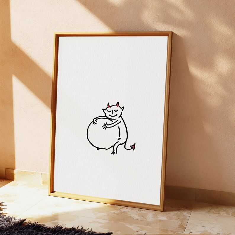 May include: A framed wall art print featuring a minimalist black line drawing of a small devil character with red horns and a red-tipped tail, hugging a large circular shape, centred on a plain white background within a light wood frame.