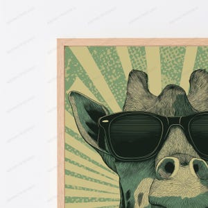 May include: A framed wall art print featuring a stylized illustration of a giraffe wearing black sunglasses. The giraffe is rendered in shades of green and tan with a sunburst background pattern. The frame is made of light-colored wood.