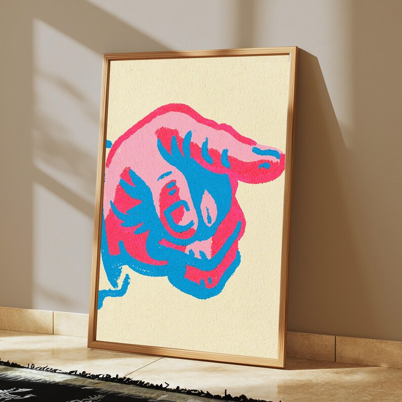 May include: A framed rectangular wall art piece featuring a stylised graphic illustration of a hand pointing to the right. The design uses bold, contrasting colours of bright pink and vibrant blue against a textured, light cream-coloured background.