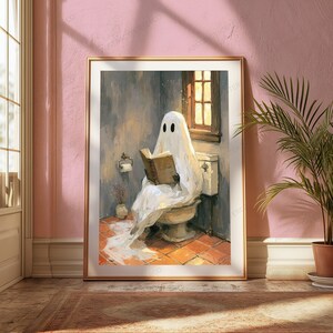 May include: A framed wall art print depicting a white ghost draped in a sheet sitting on a toilet while reading an open book. The scene features a tiled floor, a window with warm light, a toilet paper holder, and a small vase in a bathroom setting.