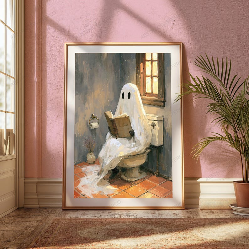 May include: A framed wall art print depicting a white ghost draped in a sheet sitting on a toilet while reading an open book. The scene features a tiled floor, a window with warm light, a toilet paper holder, and a small vase in a bathroom setting.