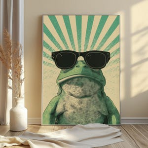 K&ouml;nnte beinhalten: Ein rechteckiger Wandkunstdruck, der einen gr&uuml;nen Frosch mit einer gro&szlig;en schwarzen Sonnenbrille zeigt. Der Hintergrund besteht aus einem cremefarbenen Feld mit strahlenden t&uuml;rkisfarbenen Sonnenstrahlen. Der Frosch ist zentriert.