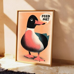 May include: A framed wall art print featuring a stylised illustration of a duck with a black head, a coral-pink chest, and dark teal wings. The duck has a speech bubble above its head containing the black text "FEED ME!" on a peach background.