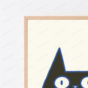 May include: A framed wall art print featuring a stylized black cat head with large white eyes and a blue outline. The cat is set against a cream-colored background, and the artwork is enclosed in a light-toned wooden frame with a clean, simple border.