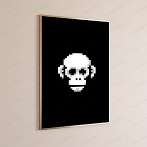 May include: A rectangular wall art print featuring a white pixelated monkey face centered on a solid black background. The artwork is encased in a thin, metallic gold frame, presented in a vertical orientation against a neutral wall surface.