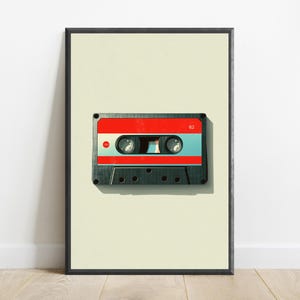 May include: A framed wall art print featuring a centered cassette tape with a black plastic shell, a bright red top section, and a light blue middle band. The number 62 is printed in white on the top right corner of the red section.