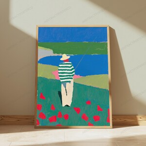 Op de afbeelding: Een ingelijste kunstdruk met een gestileerd schilderij van een persoon met een strooien hoed en een groen-wit gestreept shirt, staand in een veld met rode bloemen. De achtergrond toont een blauw wateroppervlak, groene heuvels en een blauwe lucht.