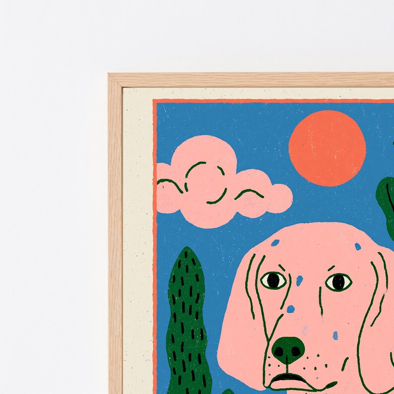 K&ouml;nnte beinhalten: Ein gerahmter Kunstdruck mit einem rosa Hund mit dunklen Flecken vor blauem Hintergrund. Die Szene zeigt eine leuchtend orangefarbene Sonne, rosa Wolken und hohe gr&uuml;ne Kakteen. Das Kunstwerk befindet sich in einem hellen Holzrahmen mit Struktur.