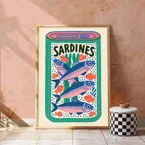May include: A framed wall art print featuring a stylized illustration of a sardine tin. The design includes three large blue and pink fish, several smaller orange fish, and green seaweed elements. The word SARDINES is printed in black at the top.