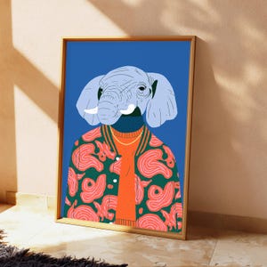 May include: A framed wall art print featuring a stylized illustration of an elephant head on a human body. The figure wears a bright orange sweater under a green varsity jacket with a pink swirling pattern, set against a solid blue background.