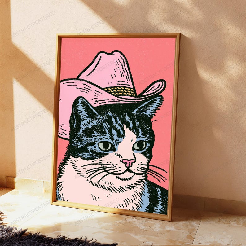 May include: A framed wall art print featuring a black and white cat wearing a pink cowboy hat against a solid pink background. The illustration uses bold lines and a graphic style to depict the cat's face, whiskers, and ears under the hat.