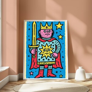 May include: A framed art print featuring a stylized illustration of a king with a beard, wearing a crown and a red cape, holding a yellow sword. The figure is set against a bright blue background decorated with yellow stars and a central sun icon.