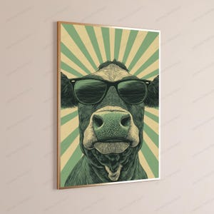 May include: A framed wall art print featuring a stylized illustration of a cow wearing black sunglasses. The cow is centered against a background of radiating light green and cream-colored sunburst stripes, presented in a vertical rectangular frame.