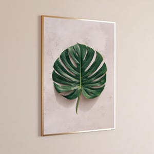 May include: A framed wall art print featuring a single, large, dark green monstera leaf centered on a light, textured, off-white background. The artwork is enclosed in a thin, metallic gold frame, creating a clean and focused botanical display.