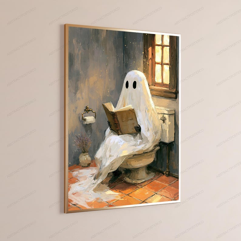 May include: A framed wall art print depicting a white ghost figure draped in a sheet, sitting on a toilet while reading an open brown book. The scene features a window with warm light, a toilet paper holder, and a small vase on a tiled floor.