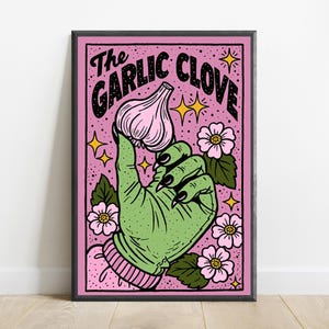 May include: A framed rectangular art print featuring a green hand with black fingernails holding a pink garlic clove. The background is pink with black stippling, surrounded by pink flowers with green leaves and yellow stars. Text reads The Garlic Clove.
