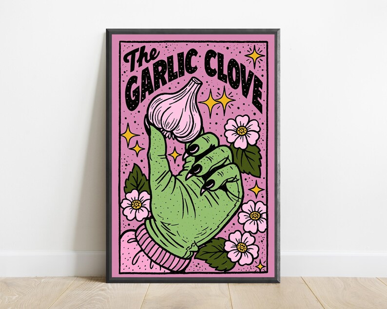 May include: A framed rectangular art print featuring a green hand with black fingernails holding a pink garlic clove. The background is pink with black stippling, surrounded by pink flowers with green leaves and yellow stars. Text reads The Garlic Clove.