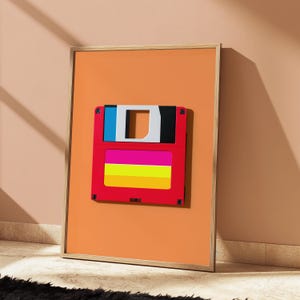 May include: A framed wall art print featuring a stylised floppy disk graphic centred on a solid orange background. The floppy disk is red with a blue and black top section and a label area displaying horizontal stripes of hot pink, yellow, and orange.