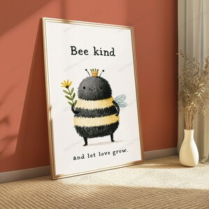 K&ouml;nnte beinhalten: Ein gerahmter Kunstdruck mit einer flauschigen, runden Hummel mit gelben und schwarzen Streifen, die eine gelbe Blume h&auml;lt. Die Hummel tr&auml;gt einen kleinen Topf auf dem Kopf. Der Text lautet Bee kind and let love grow in einfacher schwarzer Schrift.