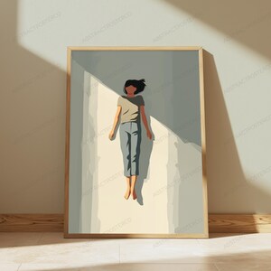 May include: A framed wall art print featuring a stylized illustration of a person floating in a light-filled space. The figure wears a beige t-shirt and blue pants, positioned against a background of soft gray and cream tones with sharp shadows.