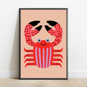 May include: A framed wall art print featuring a stylized red crab with light blue eyes and a black smile. The crab has pink spotted claws and a body with vertical purple stripes, set against a solid light peach background within a black frame.