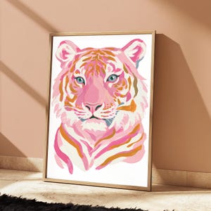 May include: A framed wall art print featuring a stylized tiger portrait. The tiger is rendered in shades of light pink, magenta, and orange, with distinct blue eyes. The artwork is set against a white background and enclosed in a light wood frame.