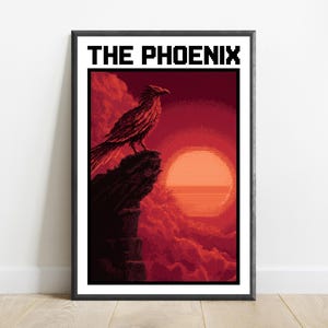 Puede incluir: Una impresi&oacute;n de arte de p&iacute;xeles enmarcada titulada THE PHOENIX, que muestra un p&aacute;jaro oscuro posado en un acantilado irregular sobre un gran sol naranja brillante contra un cielo rojo profundo y nublado. La obra est&aacute; en un marco negro.