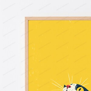 以下が含まれることがあります： 明るい黄色の背景に、下部に猫のイラストが描かれた額装されたウォールアートプリント。猫は黒と白の毛並みで、ピンクの鼻と黄色い目をしています。フレームは薄い色の木製で、細いプロファイルが特徴です。