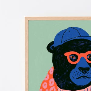 May include: A framed wall art print featuring a stylized black bear wearing a bright blue baseball cap and vibrant orange glasses. The bear is set against a solid light green background and wears a pink patterned garment around its neck.