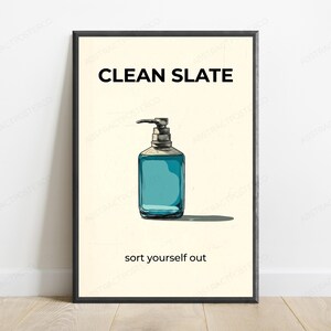 May include: A framed wall art print featuring a light cream background with a central illustration of a blue liquid soap dispenser. The text CLEAN SLATE appears in black at the top, and the text sort yourself out appears in black at the bottom.