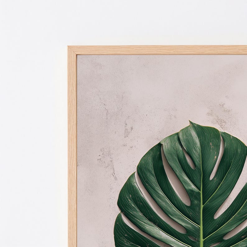 May include: A framed wall art piece featuring a large, deep green monstera leaf with characteristic splits and holes. The leaf is set against a textured, light beige or pale pinkish-grey background, enclosed within a light-toned wooden frame.