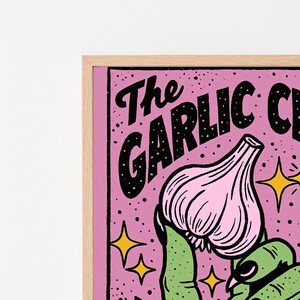 May include: A framed wall art print featuring a pink background with black speckled texture and yellow four-pointed stars. The text reads The GARLIC C in black lettering. A green hand holds a light pink garlic bulb with dark outlines.