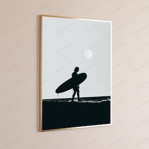 May include: A framed wall art print featuring a black and white silhouette of a person carrying a surfboard along a shoreline. The scene depicts a dark figure against a light sky with a visible moon, presented in a thin gold-colored frame.