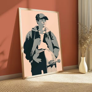 May include: A framed wall art print featuring a stylized illustration of a person wearing a baseball cap and a hooded jacket, standing while holding a skateboard. The artwork uses a palette of peach, dark gray, and light gray tones.