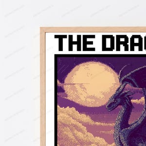 May include: A framed pixel art print featuring a dark purple dragon silhouette against a large, pale yellow moon and a cloudy sky. The text THE DRA is visible in black pixelated font at the top of the white-bordered print within a light wood frame.