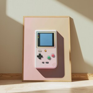 May include: A framed wall art print featuring a light pink handheld video game console with a blue screen, a black directional pad, a green button, a red button, and a small yellow button, set against a split-tone beige and pink background.