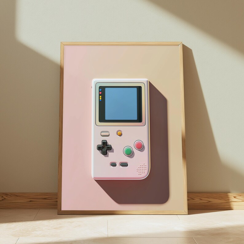 May include: A framed wall art print featuring a light pink handheld video game console with a blue screen, a black directional pad, a green button, a red button, and a small yellow button, set against a split-tone beige and pink background.