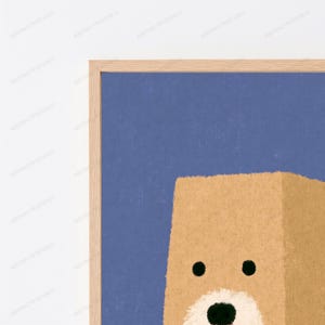 May include: A framed wall art print featuring a stylized tan bear head against a solid blue background. The bear has two small black circular eyes, a white muzzle area, and a small black nose. The frame is made of light-colored natural wood.